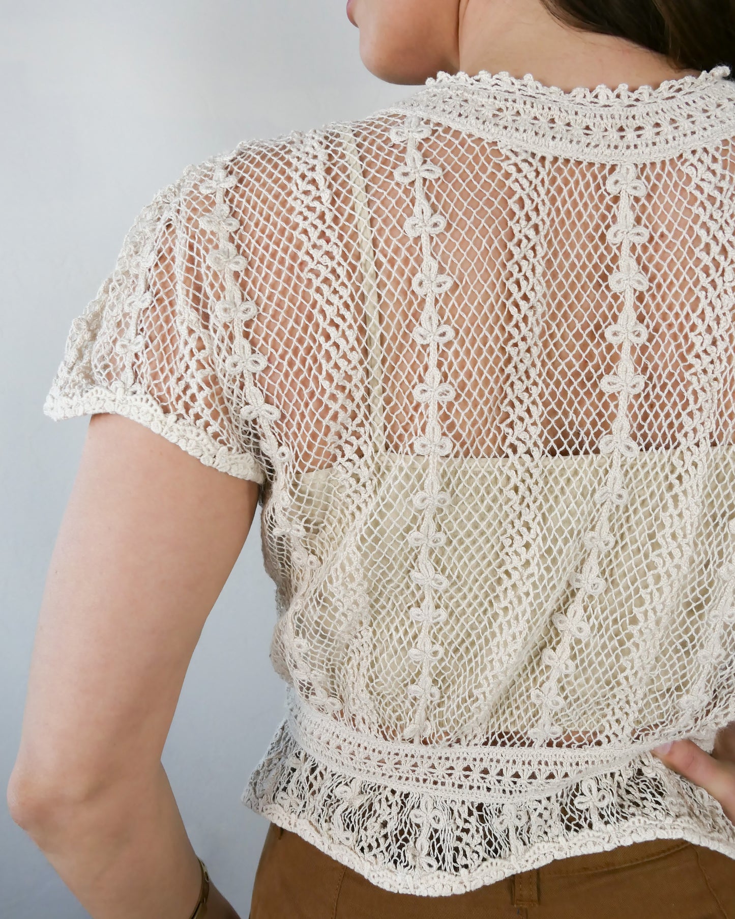 Closeup of back of top. A Lim's original vintage crochet piece from the 1980s, this top was hand crocheted with very fine threads to create a delicate, lightweight, and transparent feel. Puffed sleeves, a high waist, and flared hem lend the top a whimsical yet tailored look. The top is reversible and can be buttoned up the front or back. Natural color.