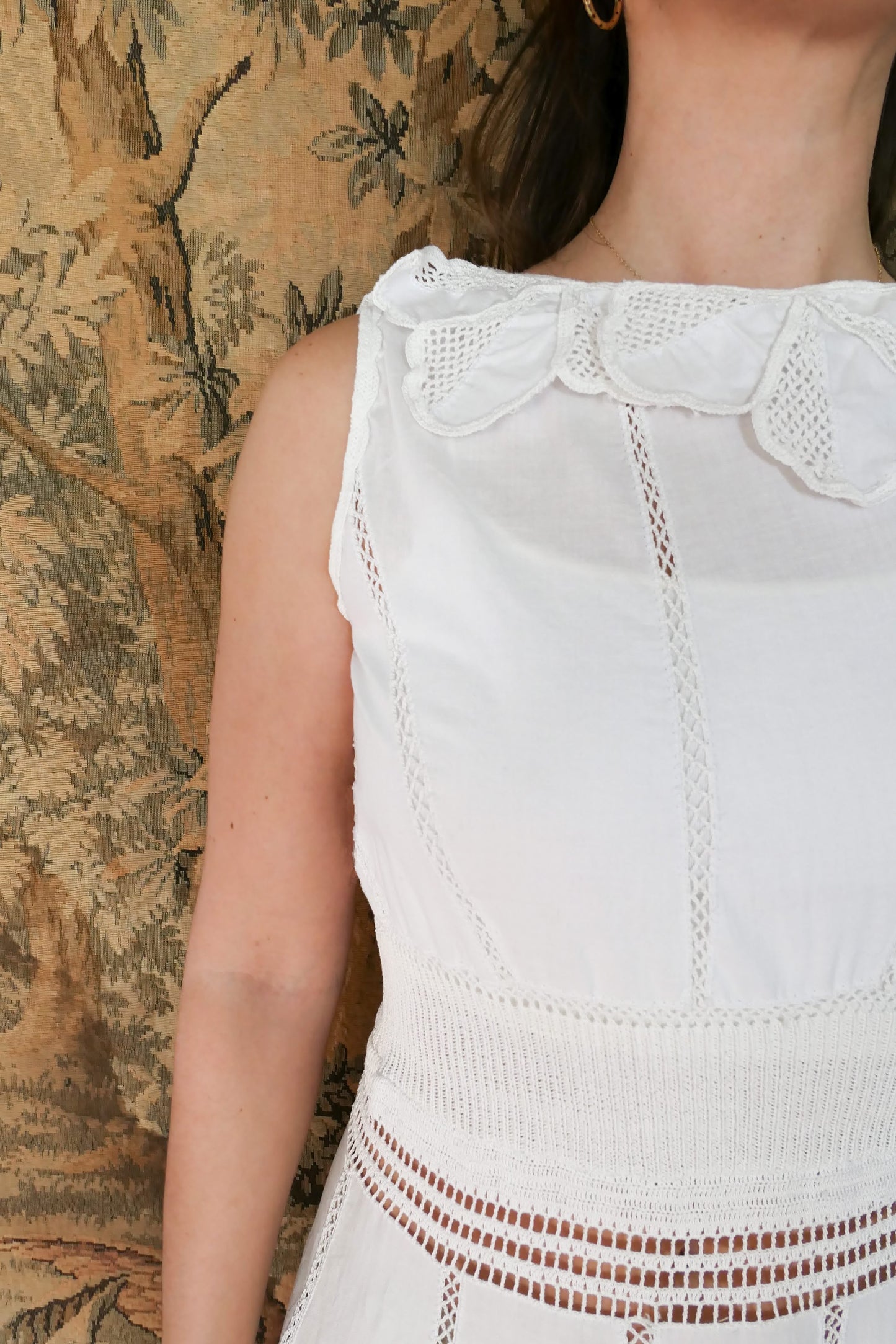 Closeup view of front of dress and collar. A white cotton midi dress with whimsical crochet and embroidered flowers at the hem and collar, and teardrop shaped eyelet detail on the skirt. Stretchable ribbed knit fabric around the waist adds contour to the dress.