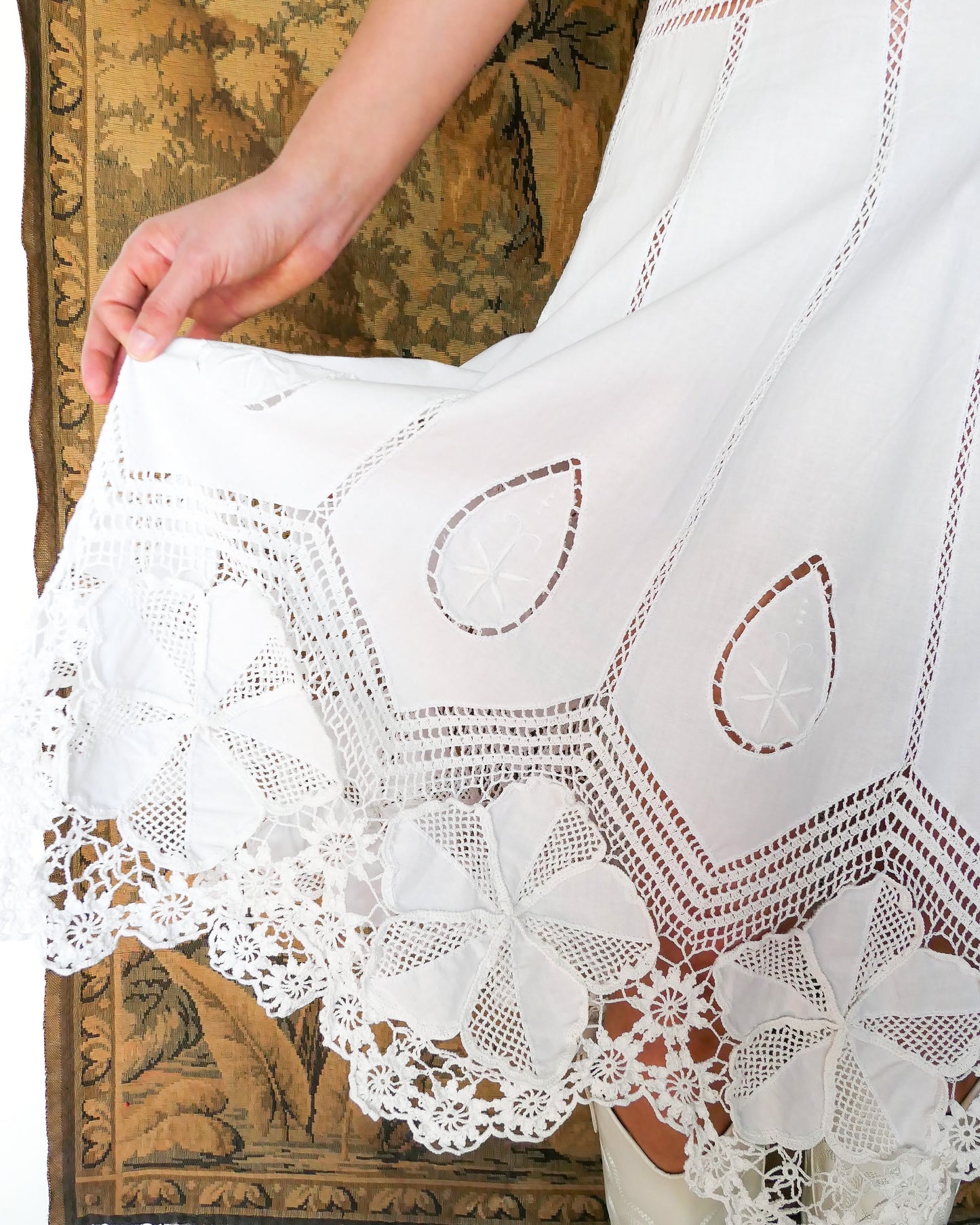 Closeup view of the hem of dress with detailed crochet and embroidery work. A white cotton midi dress with whimsical crochet and embroidered flowers at the hem and collar, and teardrop shaped eyelet detail on the skirt. Stretchable ribbed knit fabric around the waist adds contour to the dress.