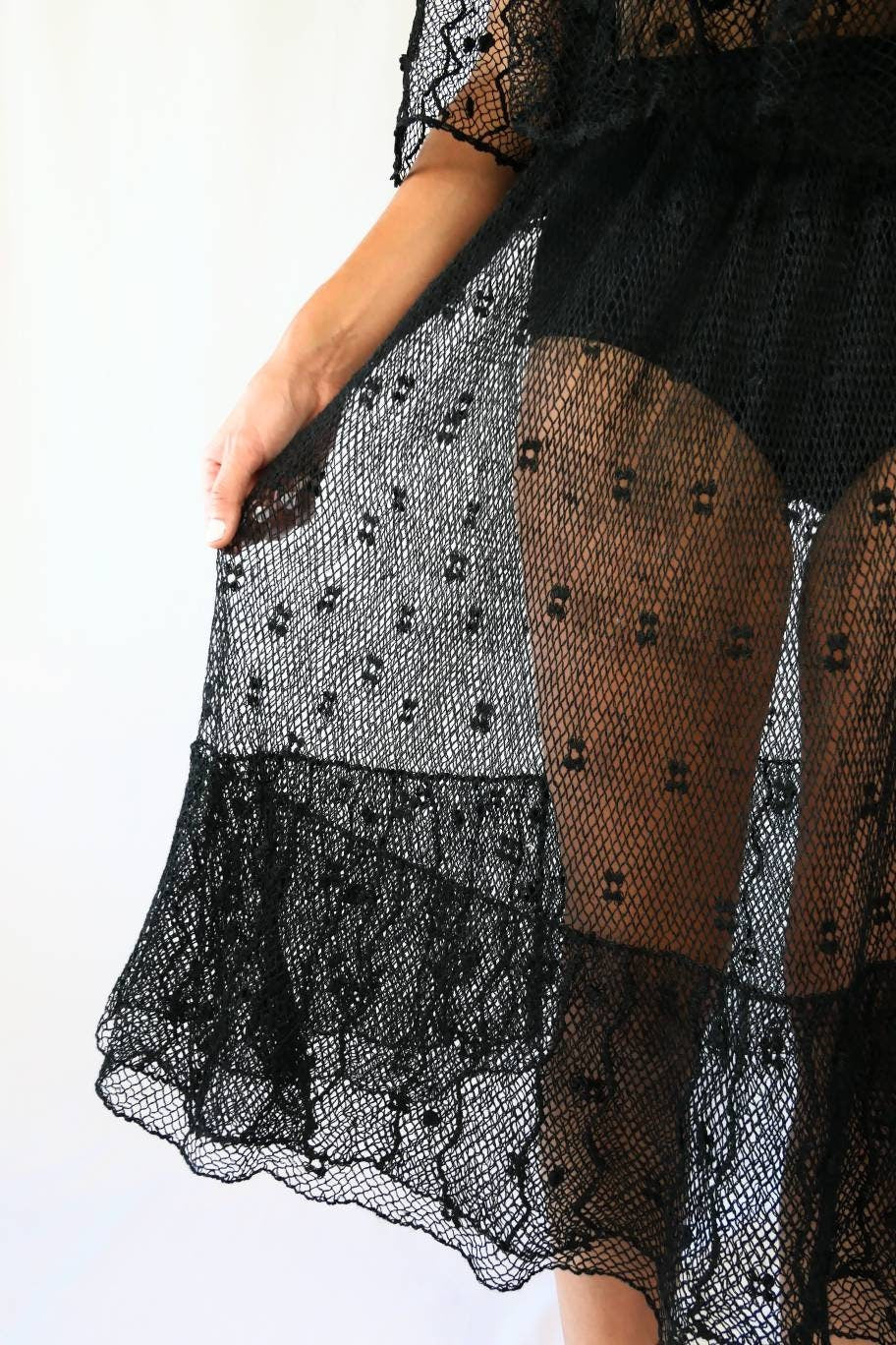 Closeup view of the bottom of dress. Sheer, delicate fishnet crochet pattern dotted with small flower-like designs. Two-tiered ruffle layers at the bottom hem.