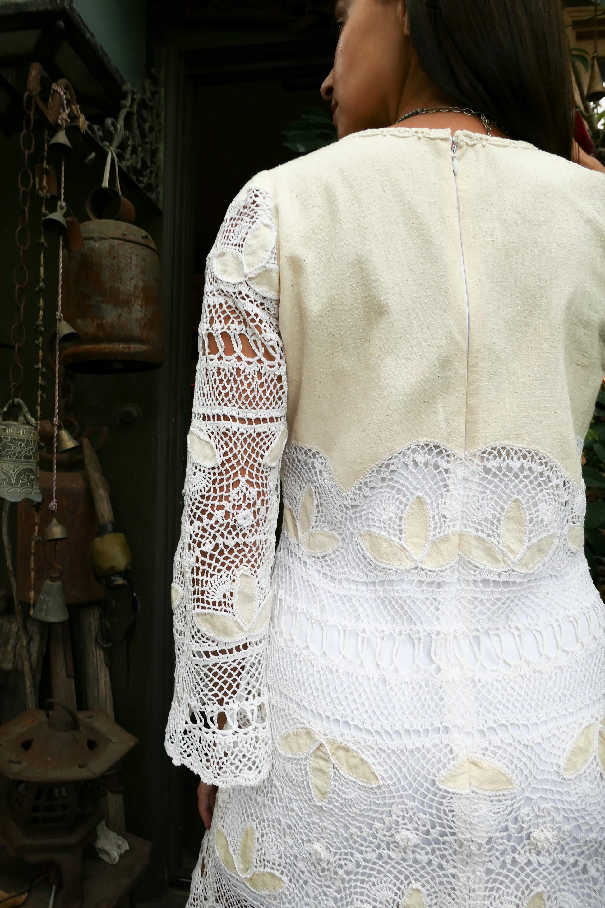 Closeup of back of dress with zipper. Natural raw silk and artisan hand crochet detail intermix in this mini dress with flared bell sleeves and a comfortable fit. This boho chic dress zips up the back, tapers in at the waist, and is perfect as a beach coverup or a highlight to your vintage wardrobe. Pair it with sandals, or sass it up with cowboy boots and a bold, vintage necklace. Colors: Natural color raw silk and white crochet