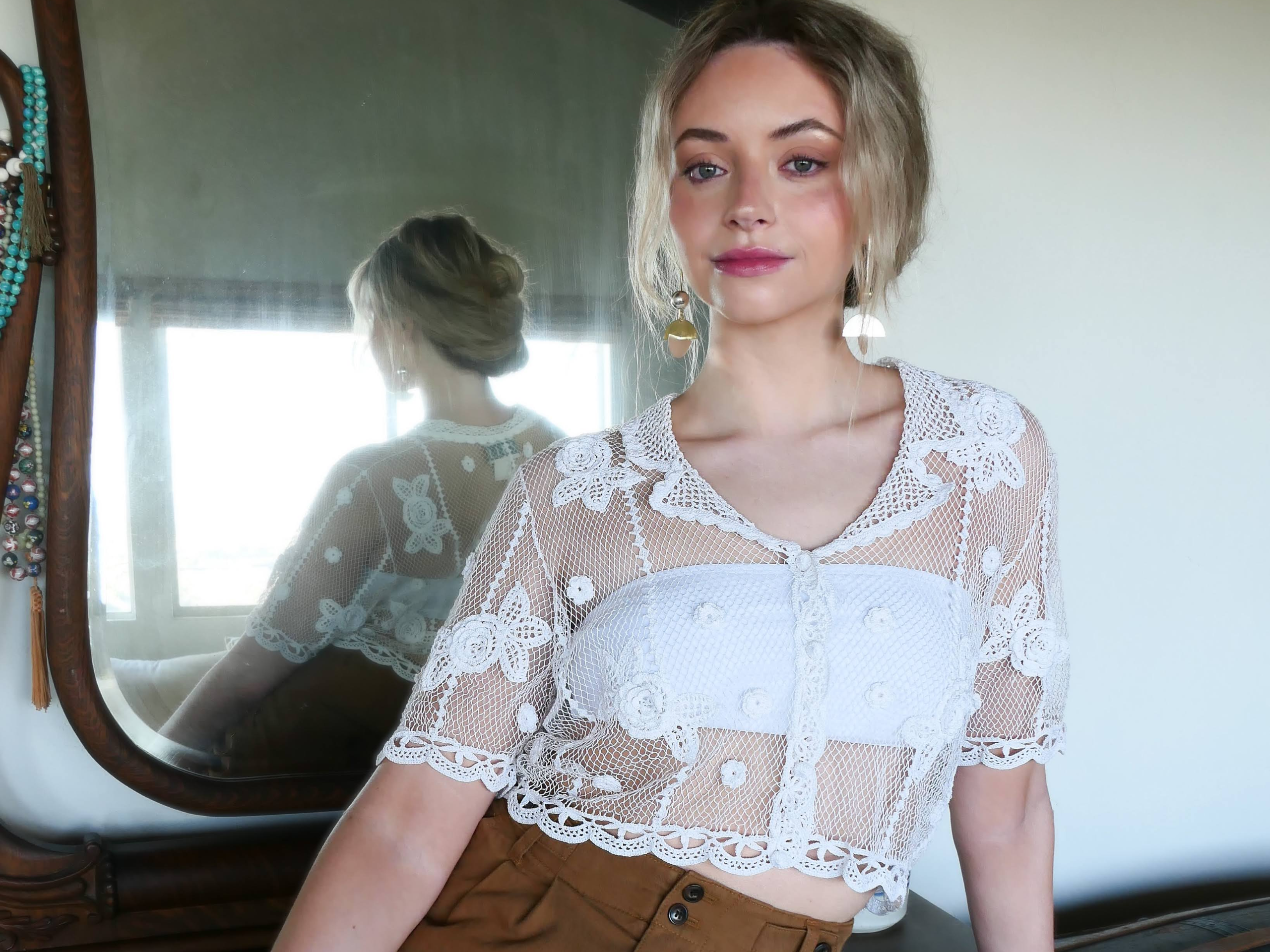 A female model is wearing a white crochet button down crop top with notched collar and carnation flowers throughout.  Scalloped trim at the sleeves and buttom hem of top.  