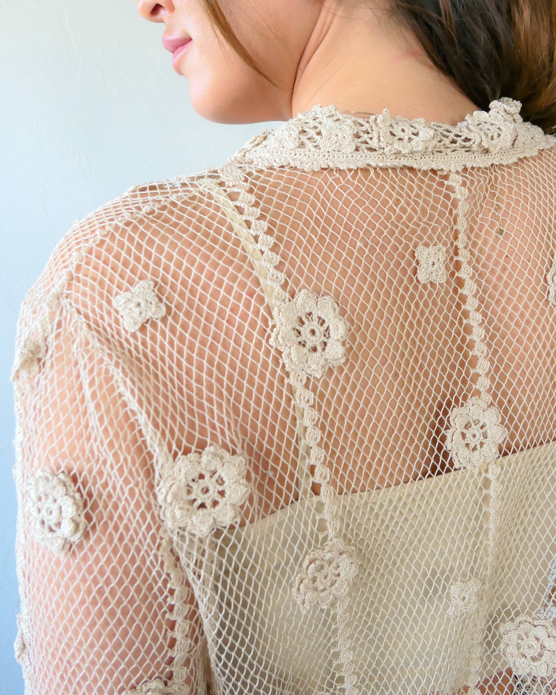 Closeup of back of top. One of the last of our original Lim's hand crocheted half sleeve button down tops in Natural from the 1990s, this versatile, lightweight, sheer top with a notched collar and floral motif design is sure to catch a few eyes. Relaxed, easy fit with dainty, hand crocheted flowers throughout and a scalloped hem reminiscent of upside down carnations. Dress it up or down any way you like.