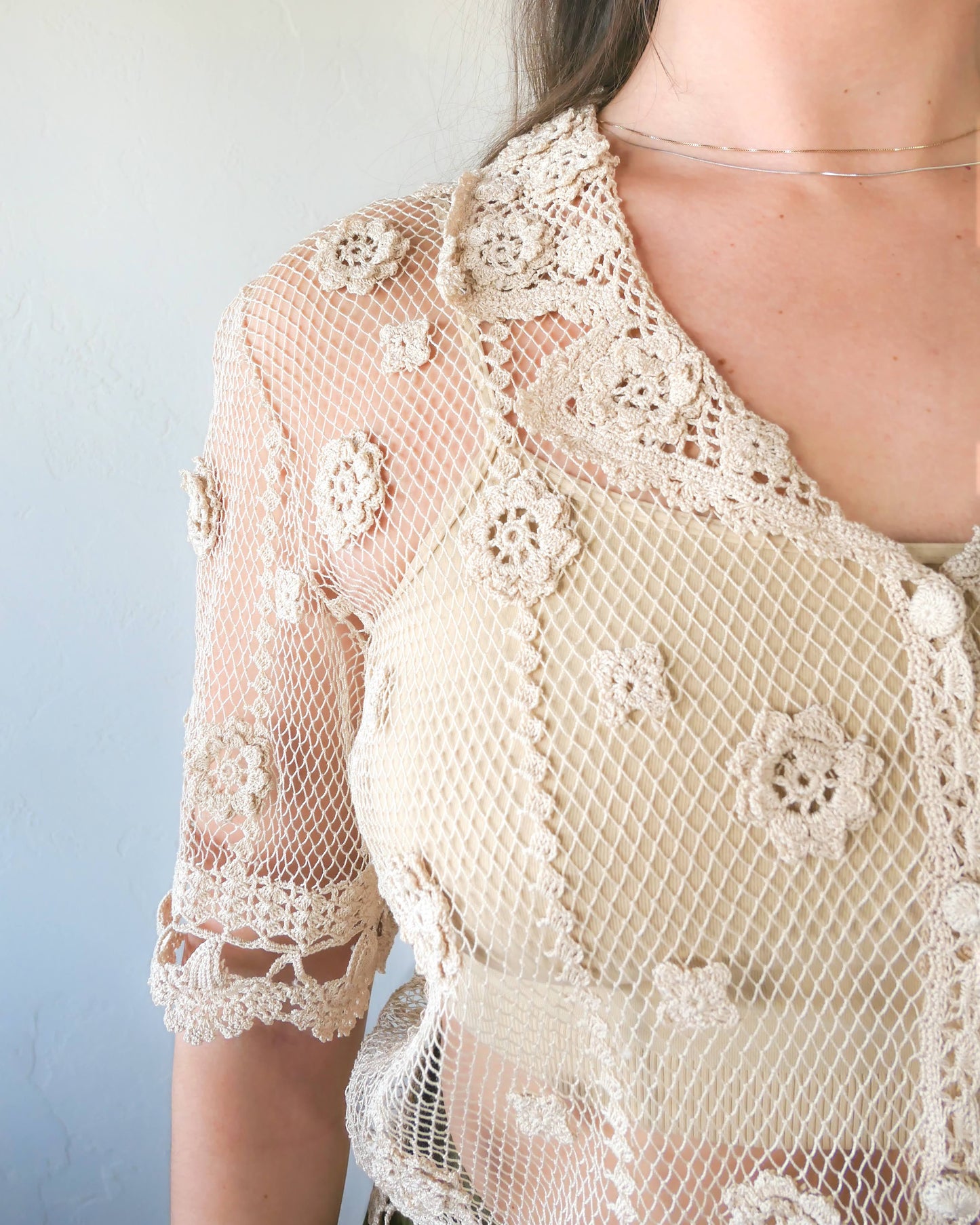 Closeup of top. One of the last of our original Lim's hand crocheted half sleeve button down tops in Natural from the 1990s, this versatile, lightweight, sheer top with a notched collar and floral motif design is sure to catch a few eyes. Relaxed, easy fit with dainty, hand crocheted flowers throughout and a scalloped hem reminiscent of upside down carnations. Dress it up or down any way you like.