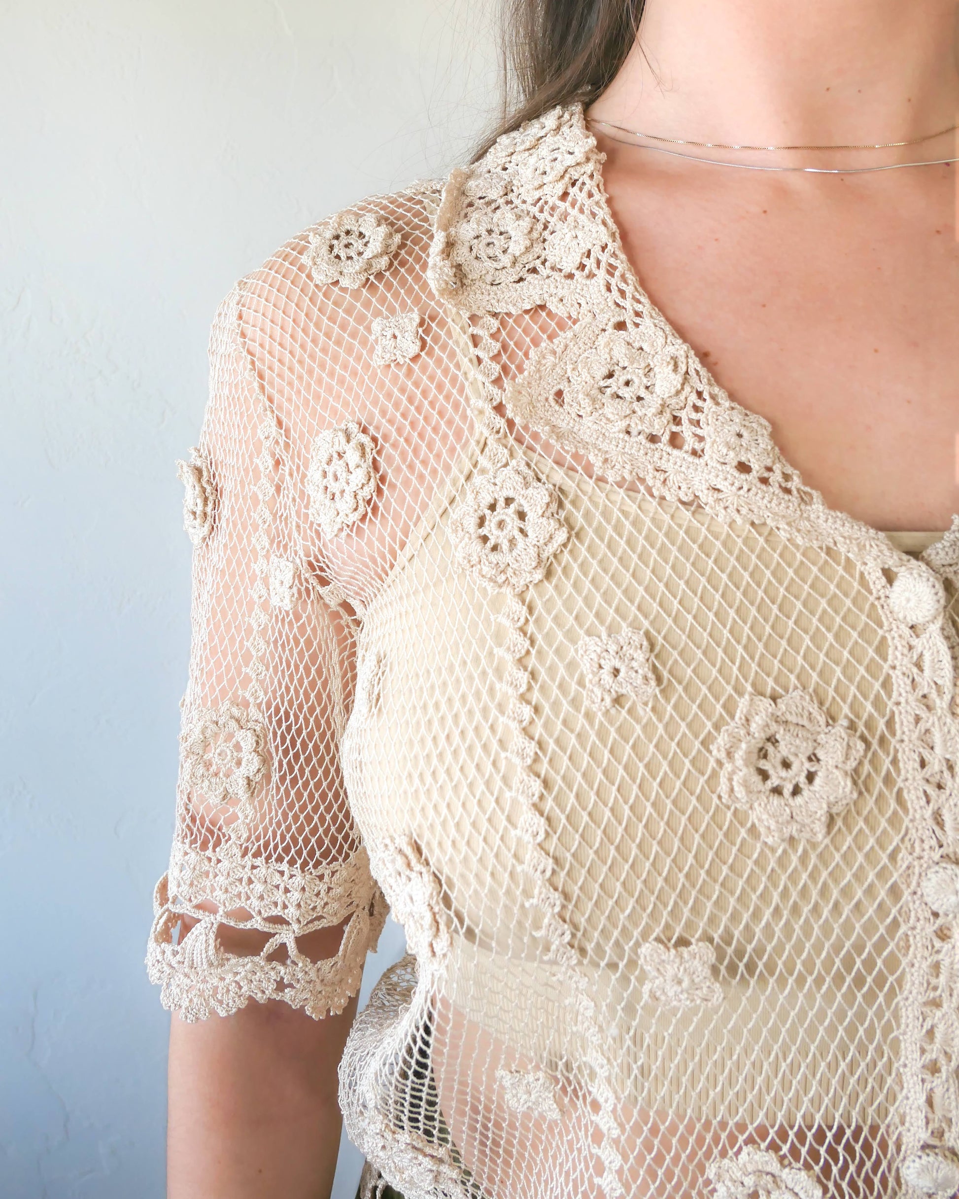 Closeup of top. One of the last of our original Lim's hand crocheted half sleeve button down tops in Natural from the 1990s, this versatile, lightweight, sheer top with a notched collar and floral motif design is sure to catch a few eyes. Relaxed, easy fit with dainty, hand crocheted flowers throughout and a scalloped hem reminiscent of upside down carnations. Dress it up or down any way you like.