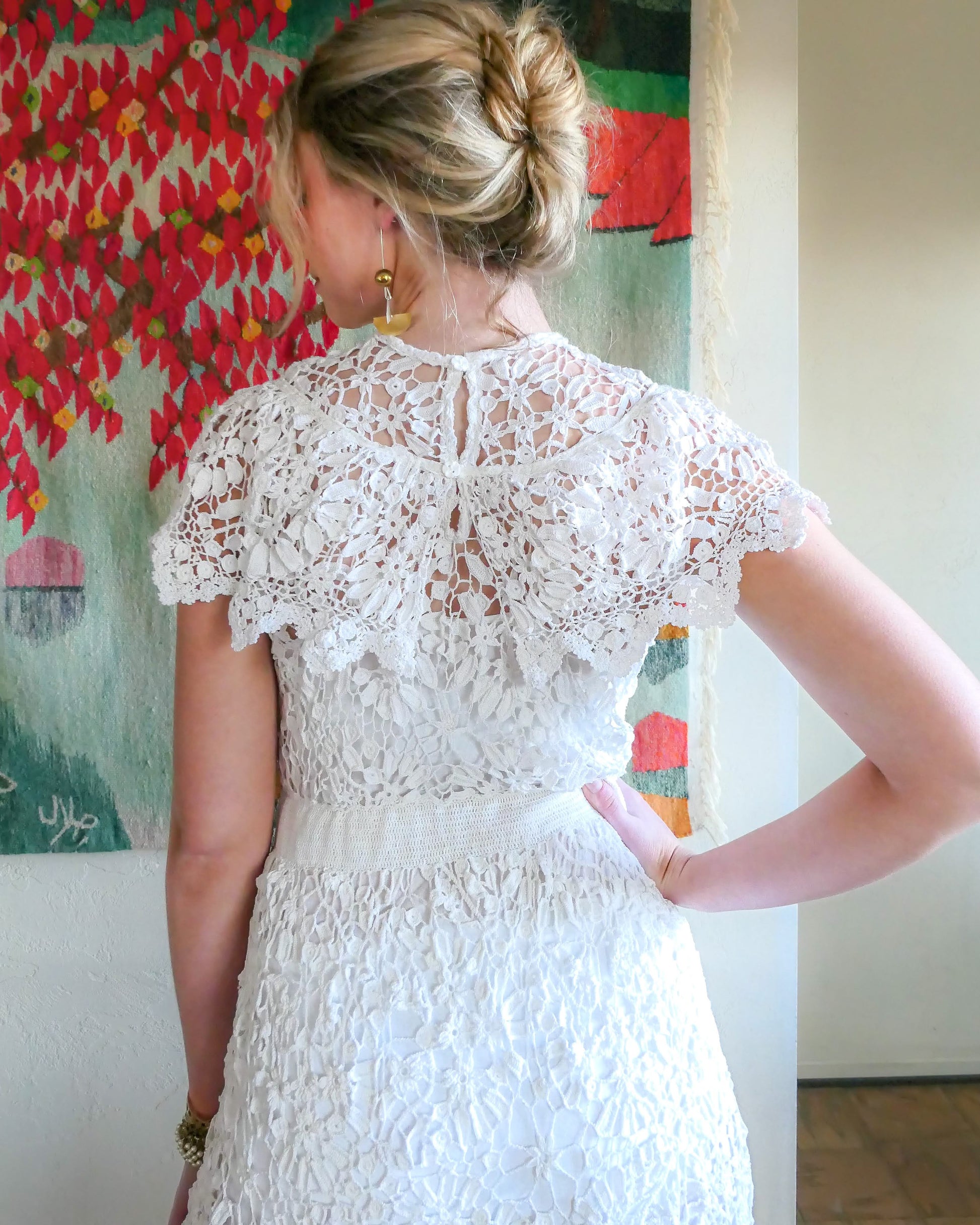 Back closeup view of dress. A romantic white crochet midi dress with a floral and leaf pattern, comfortable elastic waistband, and a flattering neck and shoulder design. Perfect as a vintage wedding dress or a special occasion! Wear with a white slip and heels for a refined, feminine look, or pair with boots for a more casual outing.