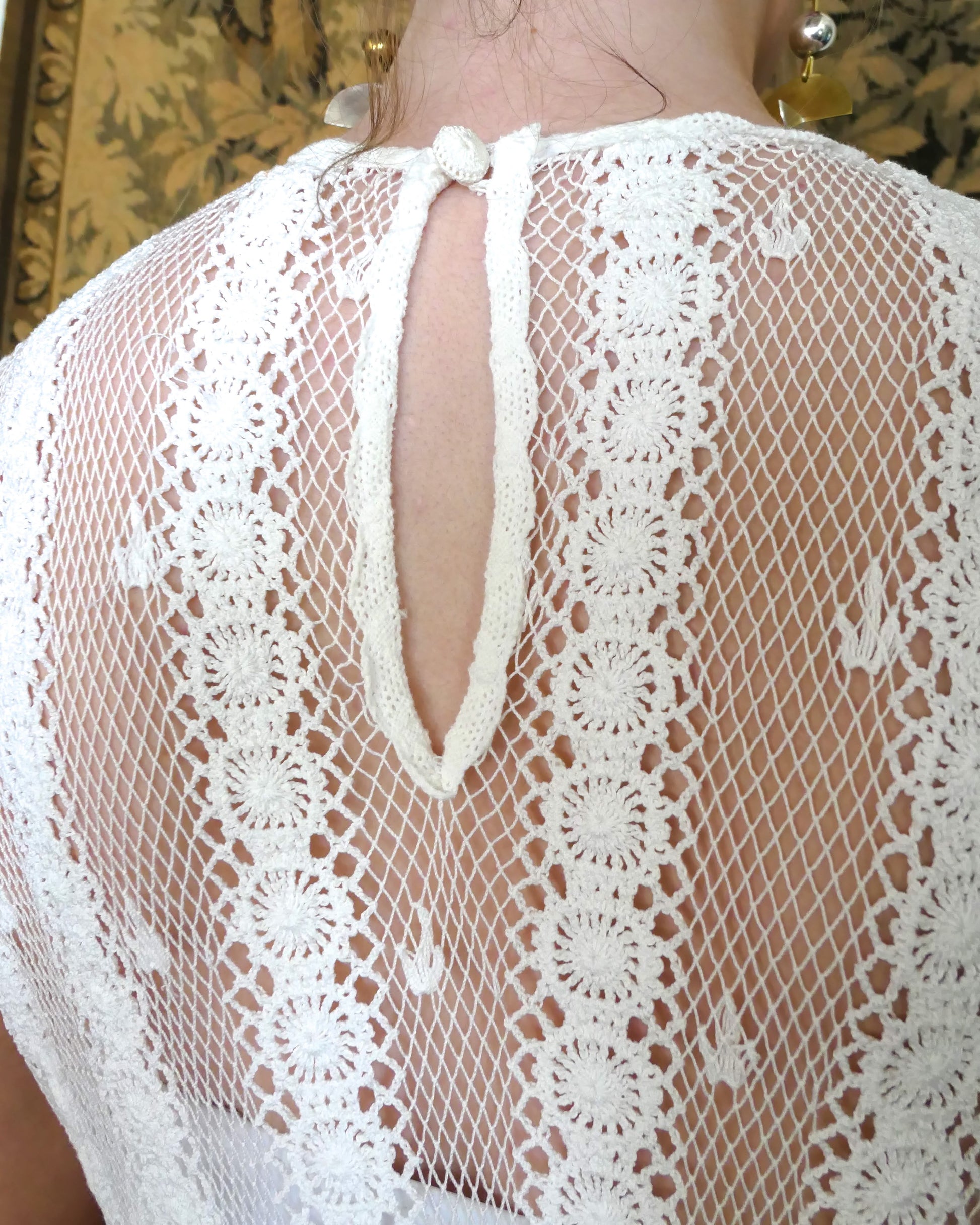 Closeup view of back of dress.  Single button at the back.  A delicate mesh crochet sleeveless midi dress in white using our vintage Lim's crochet fabric with a comfortable fitting, elastic high waist and vertical stripes  The dress flares out gradually towards the two-tiered hem and is accented with a floral trim.