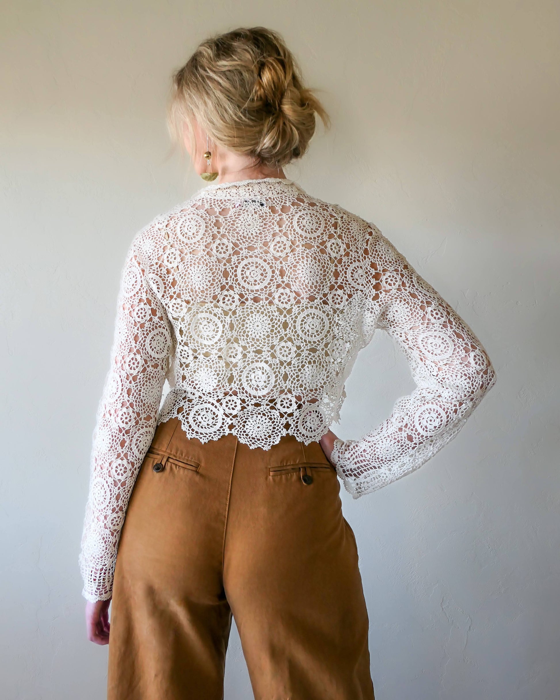Back view. A versatile and boho chic hand crocheted cropped cardigan sweater in Ivory with a unique, repeating kaleidoscope-like floral pattern throughout. Dress it down with a pair of jeans and sandals, or dress it up with neutral colored wide leg pants, heels, and a pair of vintage dangly earrings.  Size: Fits Small to Medium